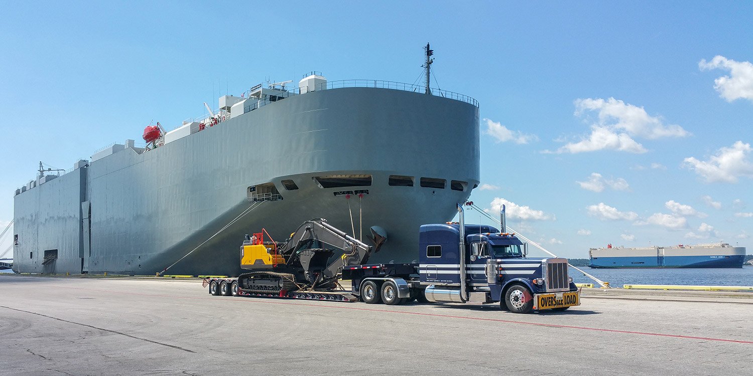 Blue Peterbilt semi with black RGN trailer parked alongside a breakbulk vessel