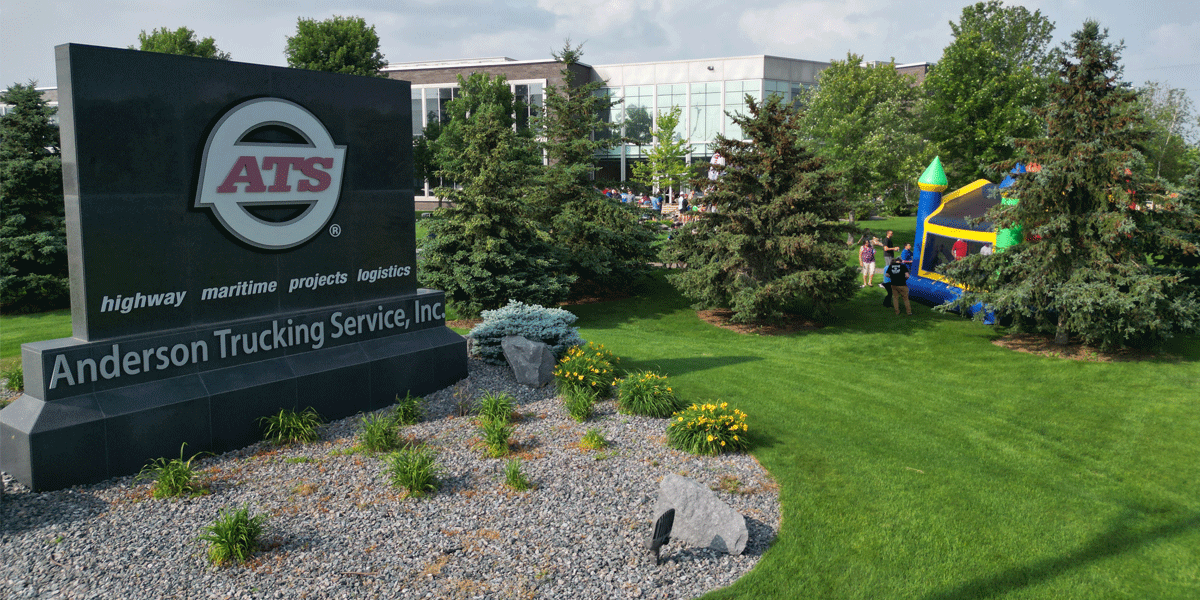 ATS' logo and slogan in granite in front of trees and bushes. A building is in the background.