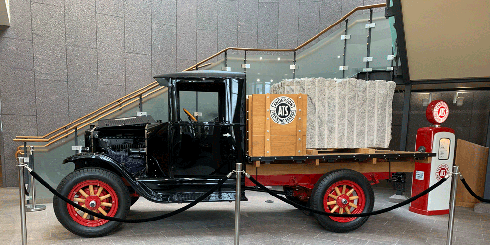 Old black truck with a large slab of granite on its bed. It's roped off in a lobby.