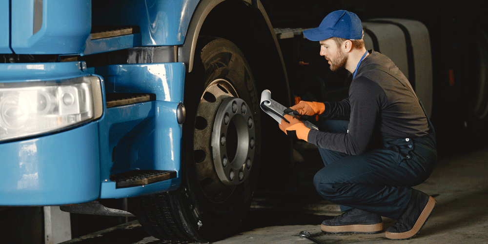 Man checking wheel and tires of a semi-truck. He holds a clipboard. 