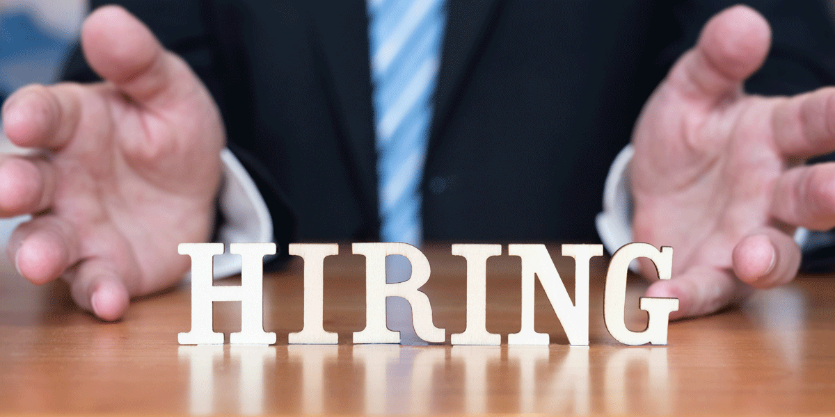 Man in suit holding his hands open in a welcoming gesture. The word hiring is in white in front of him.
