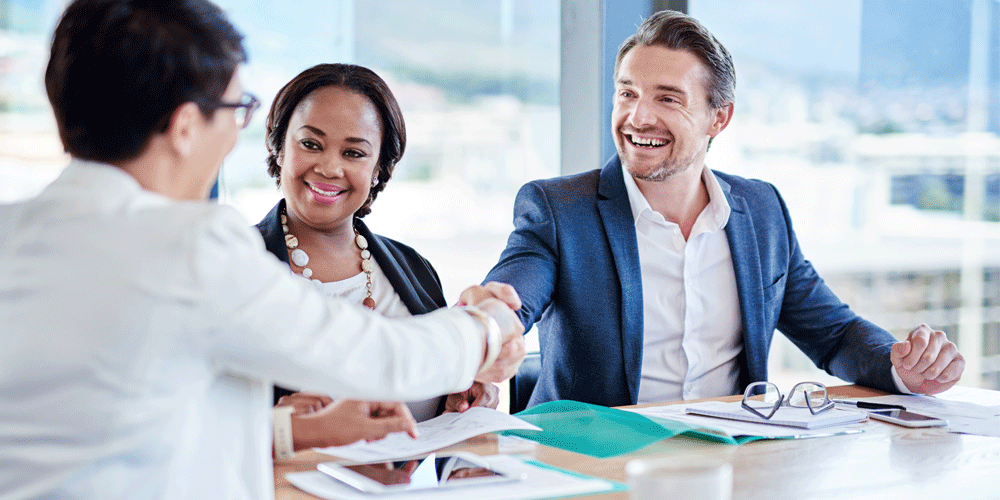 Three people sitting at a table in full-window conference room. Two of the men shake hands as they smile.