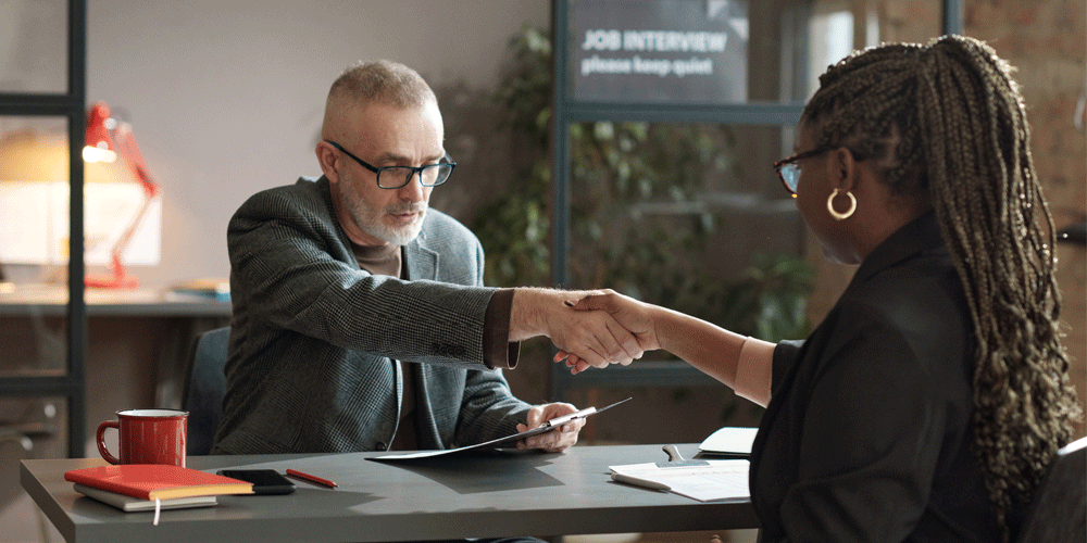 A manager shaking a job applicant's hand. They sit at a desk.