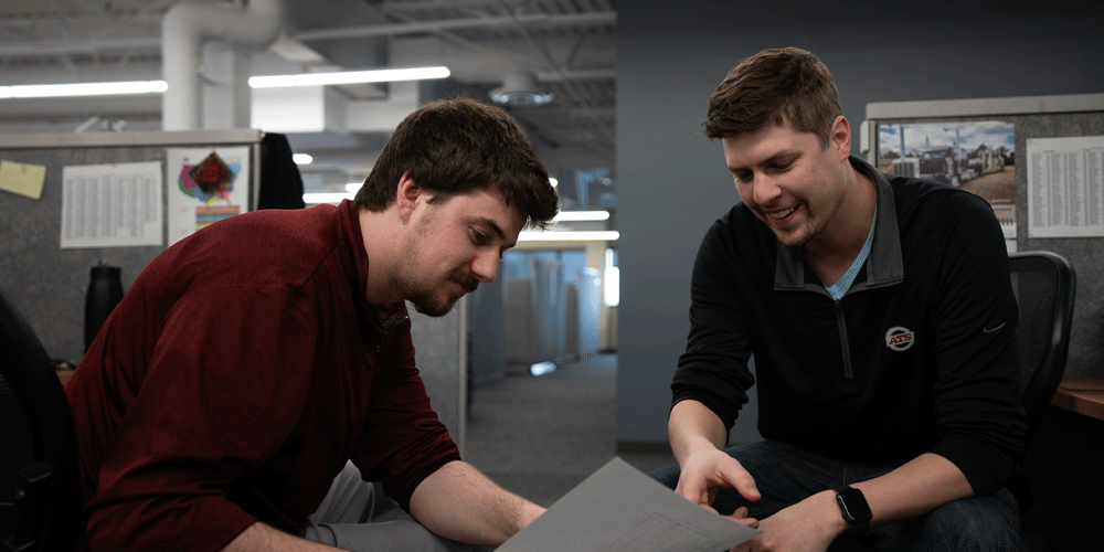 Two men sitting between cubicles looking at a piece of paper.